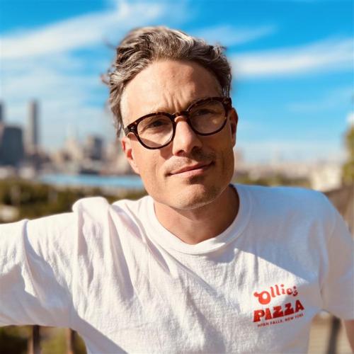 Headshot of man wearing a white t-shirt and black glasses in standing in front of a city skyline