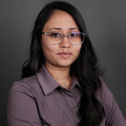 Headshot of a woman with long dark hair, glasses and wearing a purple shirt in front of a dark grey background