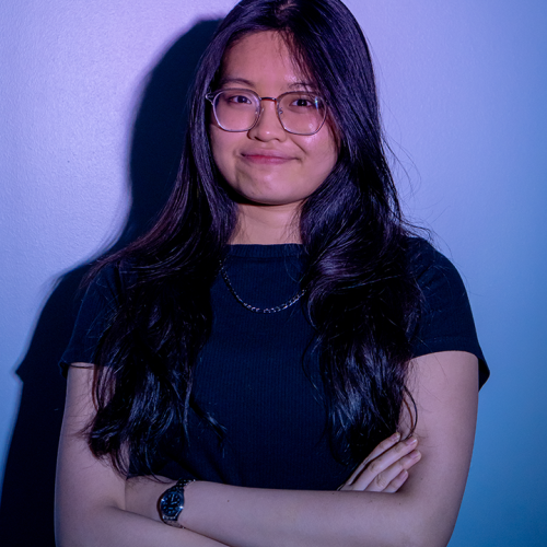 headshot of a girl with glasses and dark long hair wearing black shirt and standing in front of a blue background
