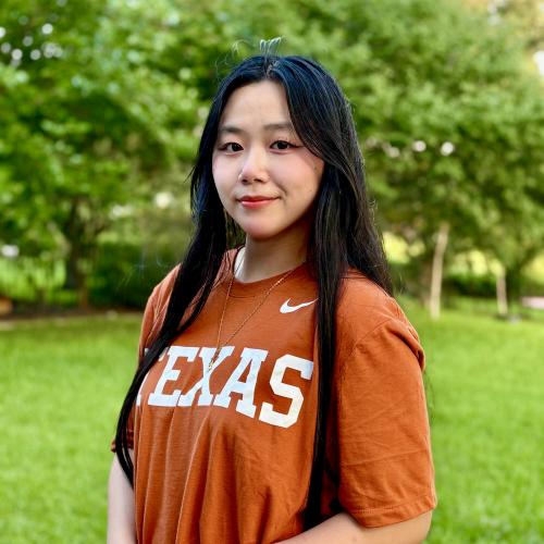 Headshot of a woman with long dark hair wearing a burnt orange shirt with a tree in the background